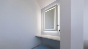 Pequeño espacio cerrado con un banco, un espejo con iluminación y una puerta.
