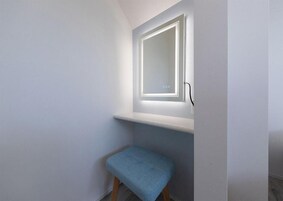 Pequeño espacio cerrado con un banco, un espejo con iluminación y una puerta.