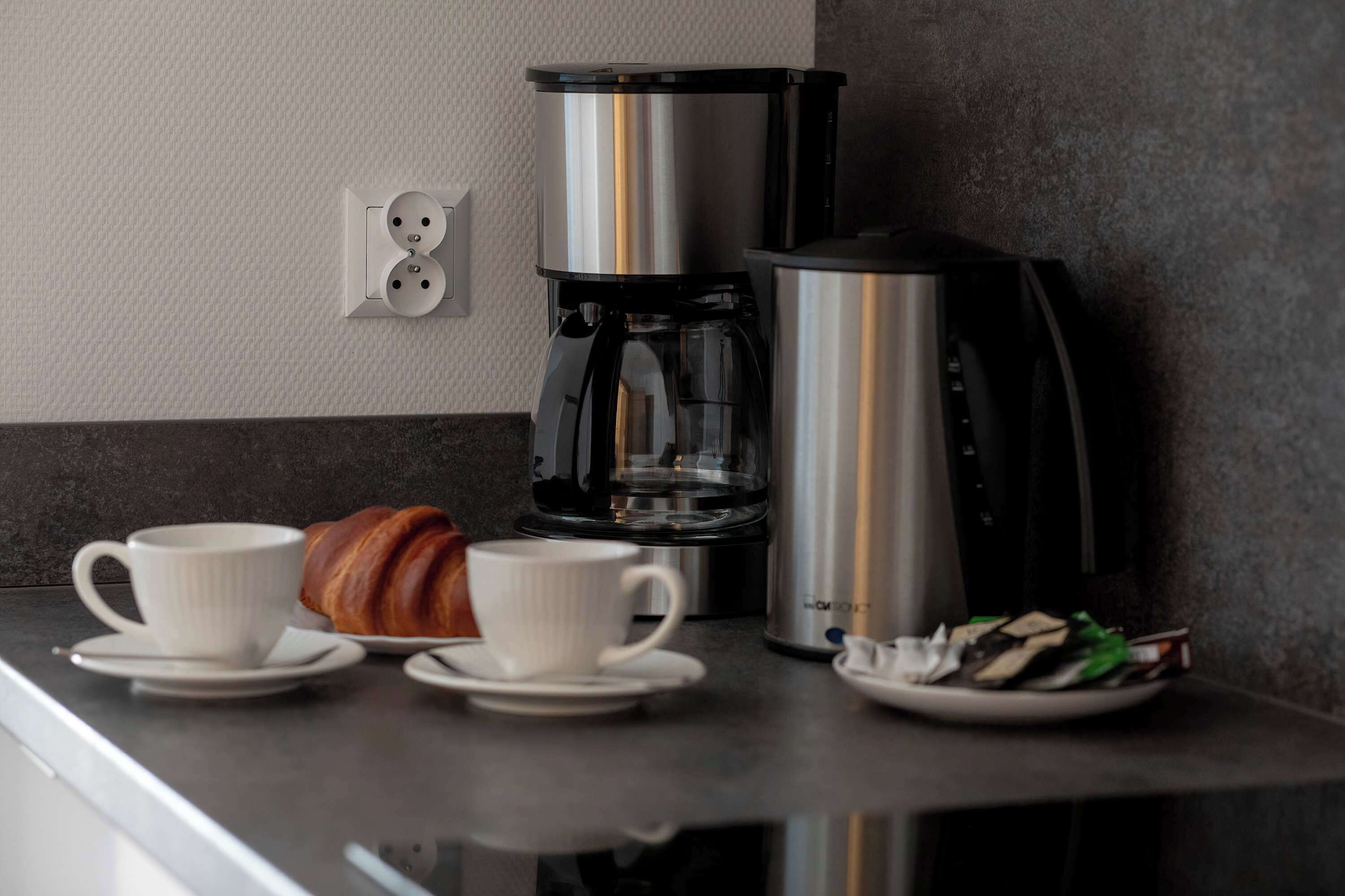 Une cuisine moderne équipée d’une machine à café, de deux tasses, d’un croissant et d’une assiette de sachets de thé.