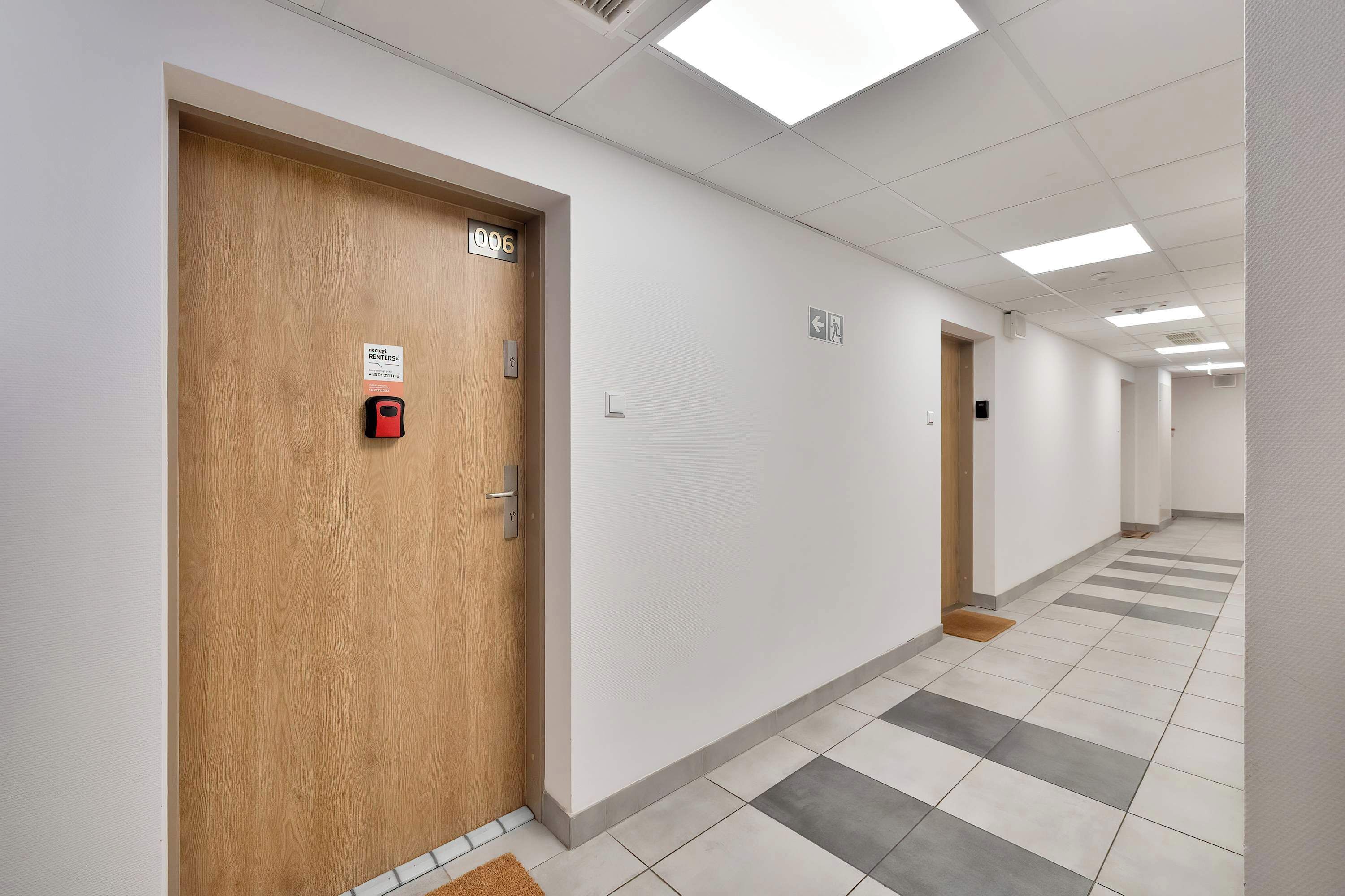 Un couloir avec des portes en bois, des panneaux numérotés et un système d’alarme incendie.