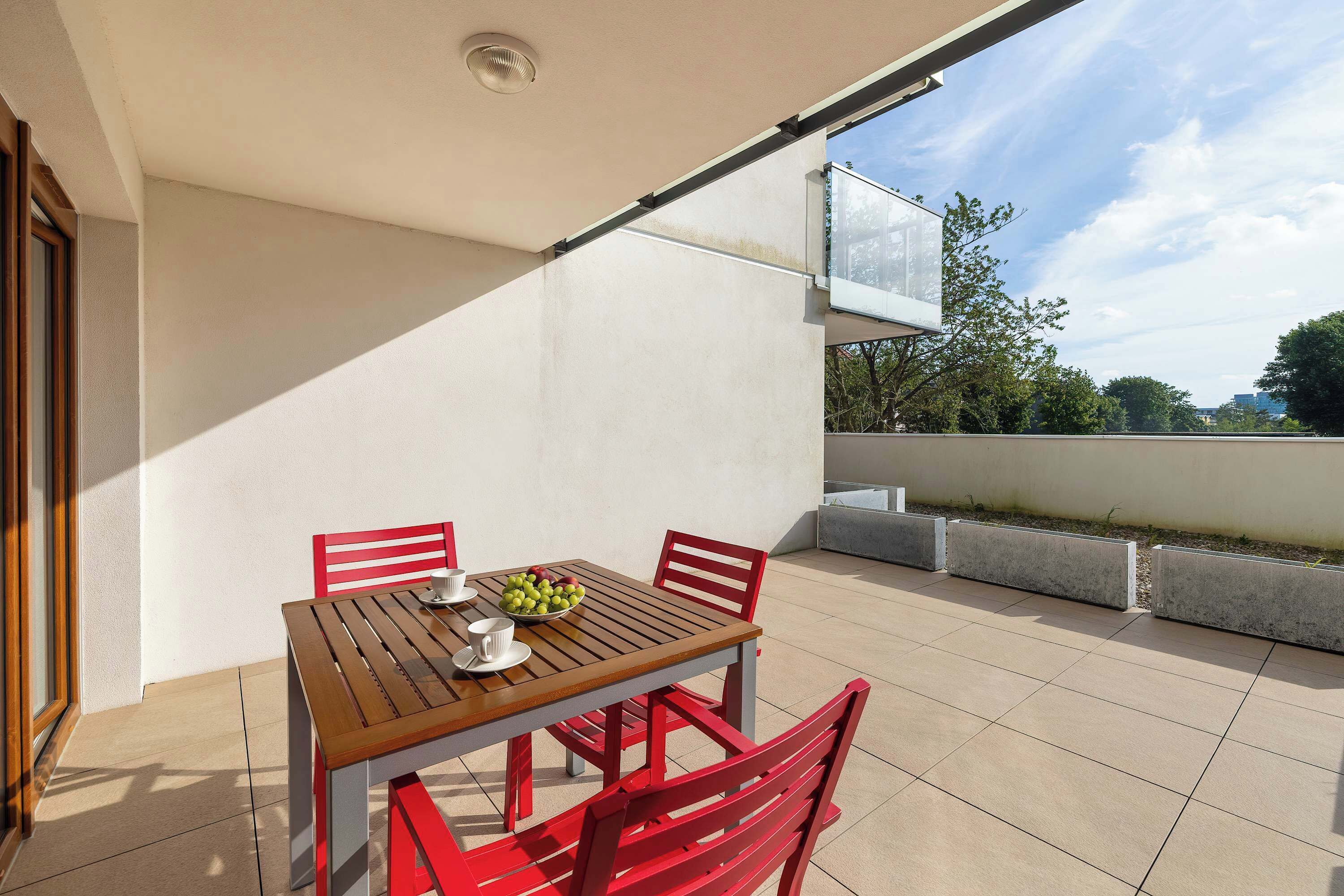 Un balcon avec une table en bois et des chaises rouges, un saladier rempli de fruits et une vue sur le paysage urbain.