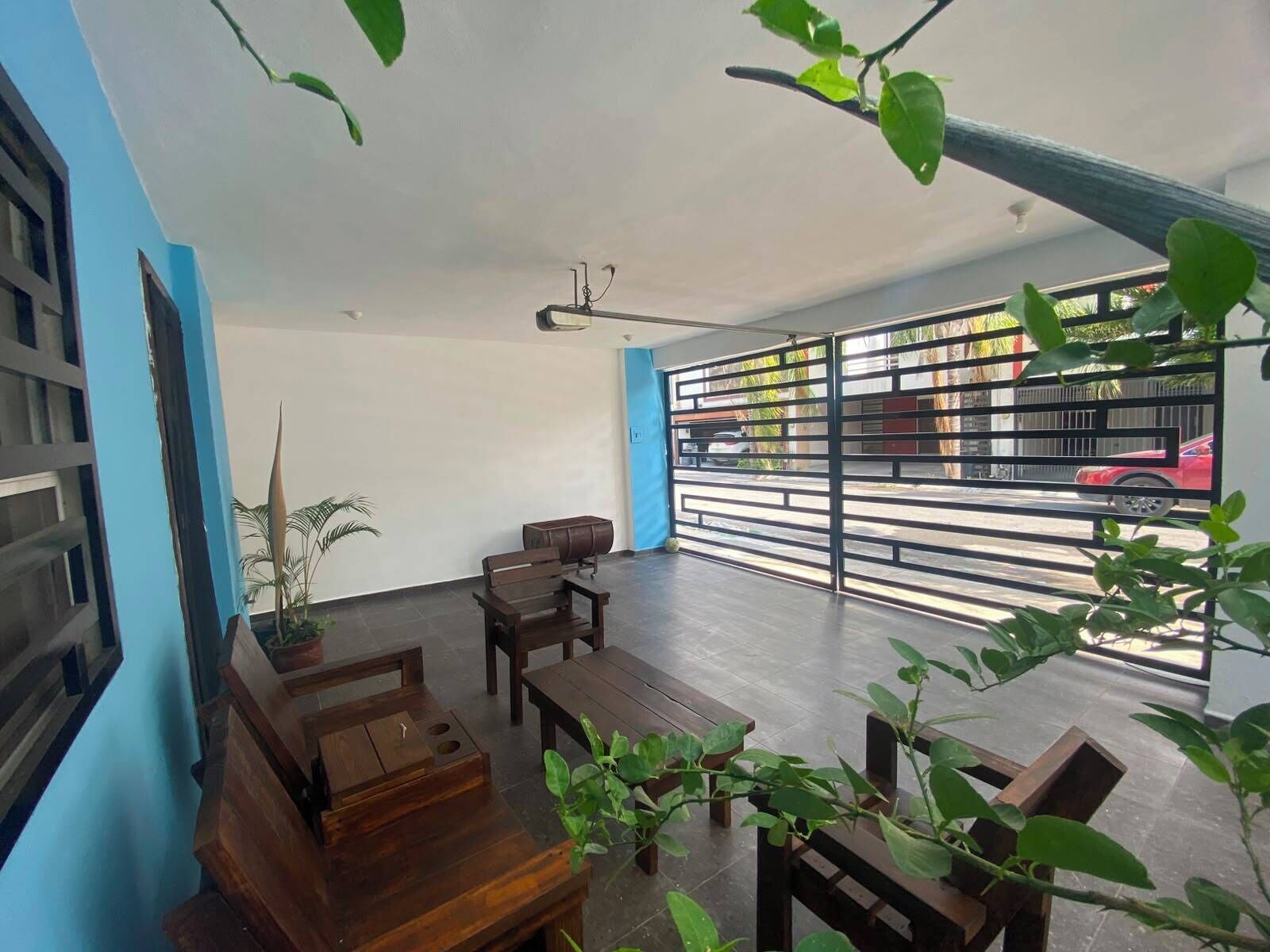 An indoor space with a wooden table and chairs, a potted plant, and a view through a metal gate to the outside.
