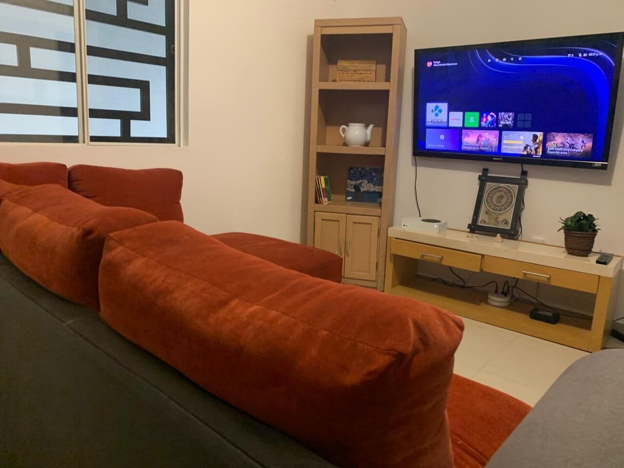 A living room with a sofa, a television mounted on the wall, a cabinet, and a window with a geometric pattern.