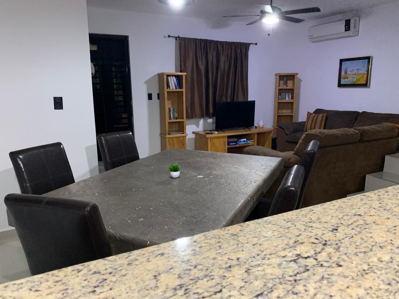 A dining area with a granite table, black chairs, a small potted plant, and a television on a wooden stand.