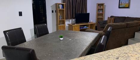 A dining area with a granite table, black chairs, a small potted plant, and a television on a wooden stand.