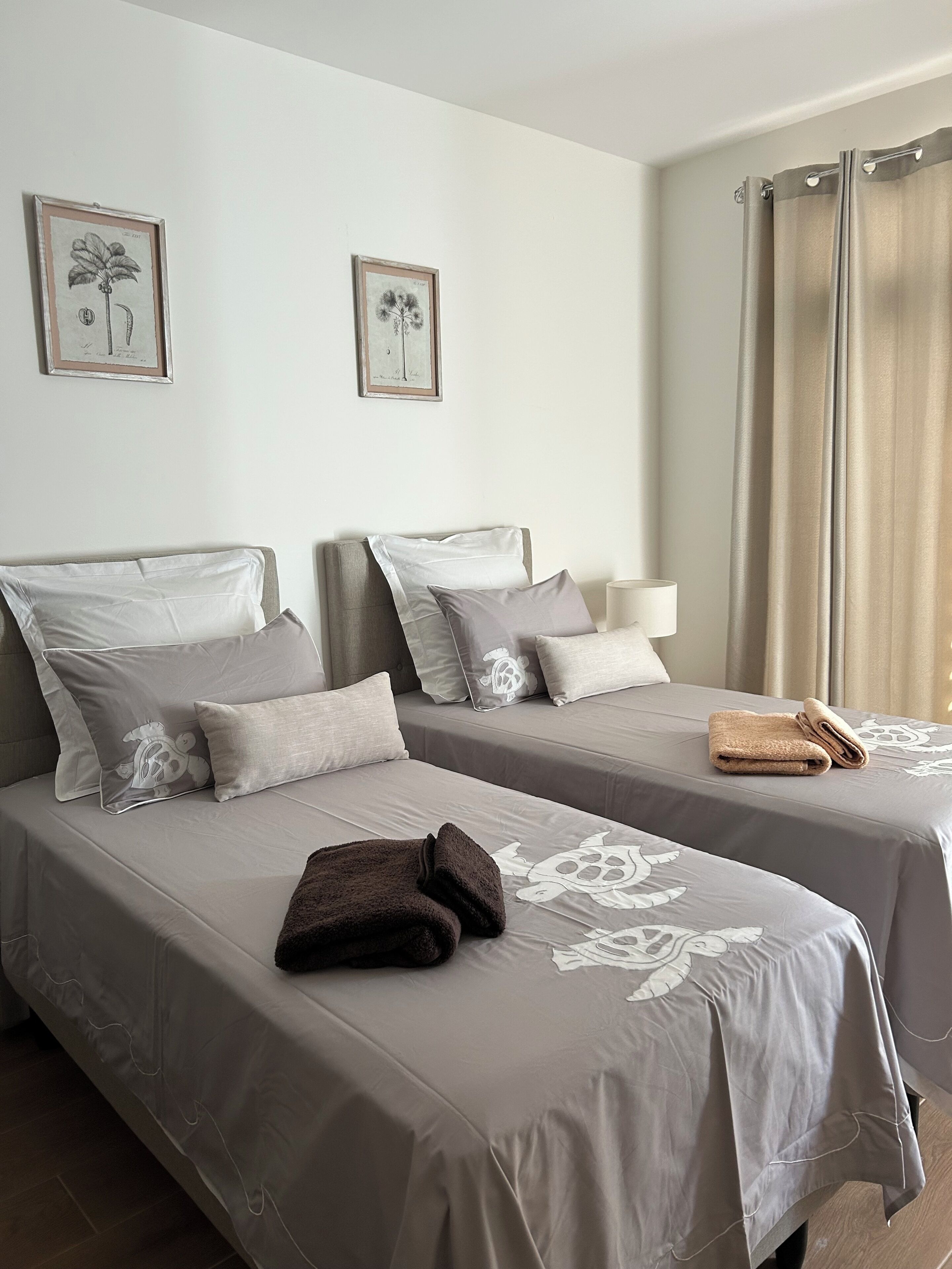 Une chambre d’hôtel avec deux lits, chacun recouvert d’une couvre-lit grise et agrémenté d’oreillers assortis, une lampe de chevet et des reproductions encadrées de motifs botaniques accrochées au mur.