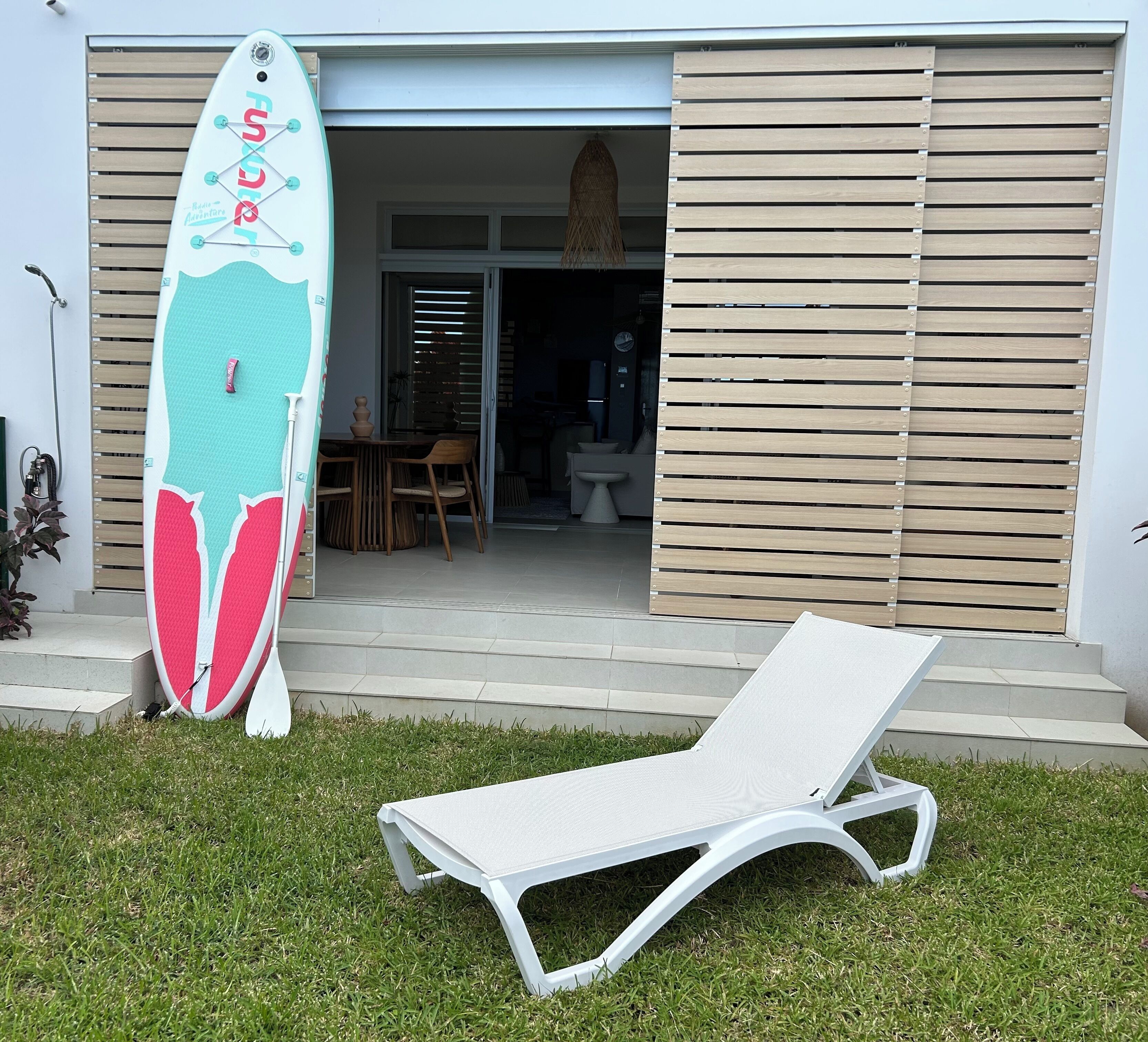 A modern house with a white lounge chair in the front yard, a surfboard leaning against the house, and a wooden door.