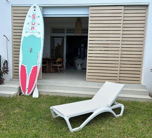 A modern house with a white lounge chair in the front yard, a surfboard leaning against the house, and a wooden door.