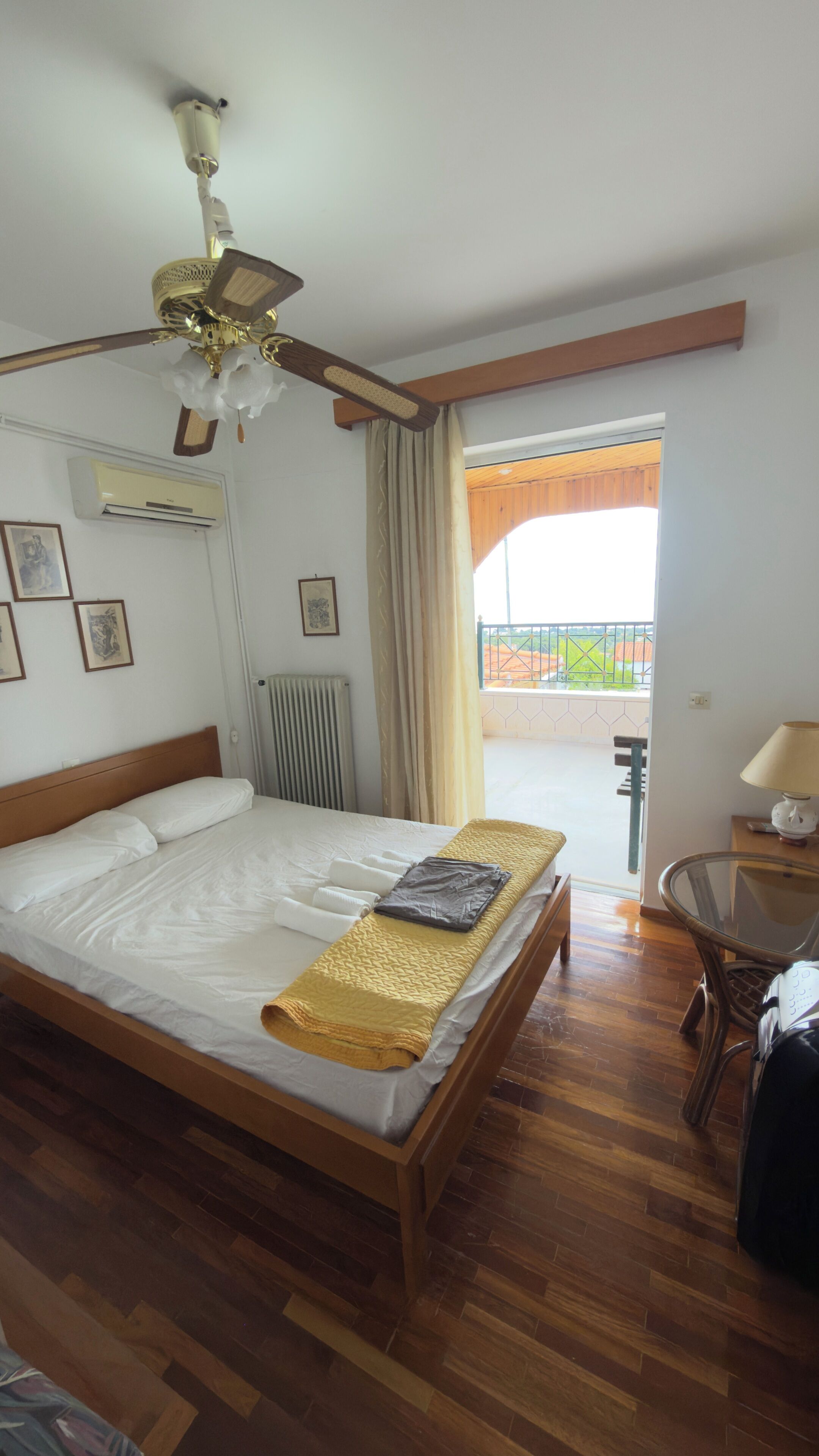 Quarto de hotel com cama, ventoinha no teto, radiador, janela com cortinas, varanda e uma cadeira.