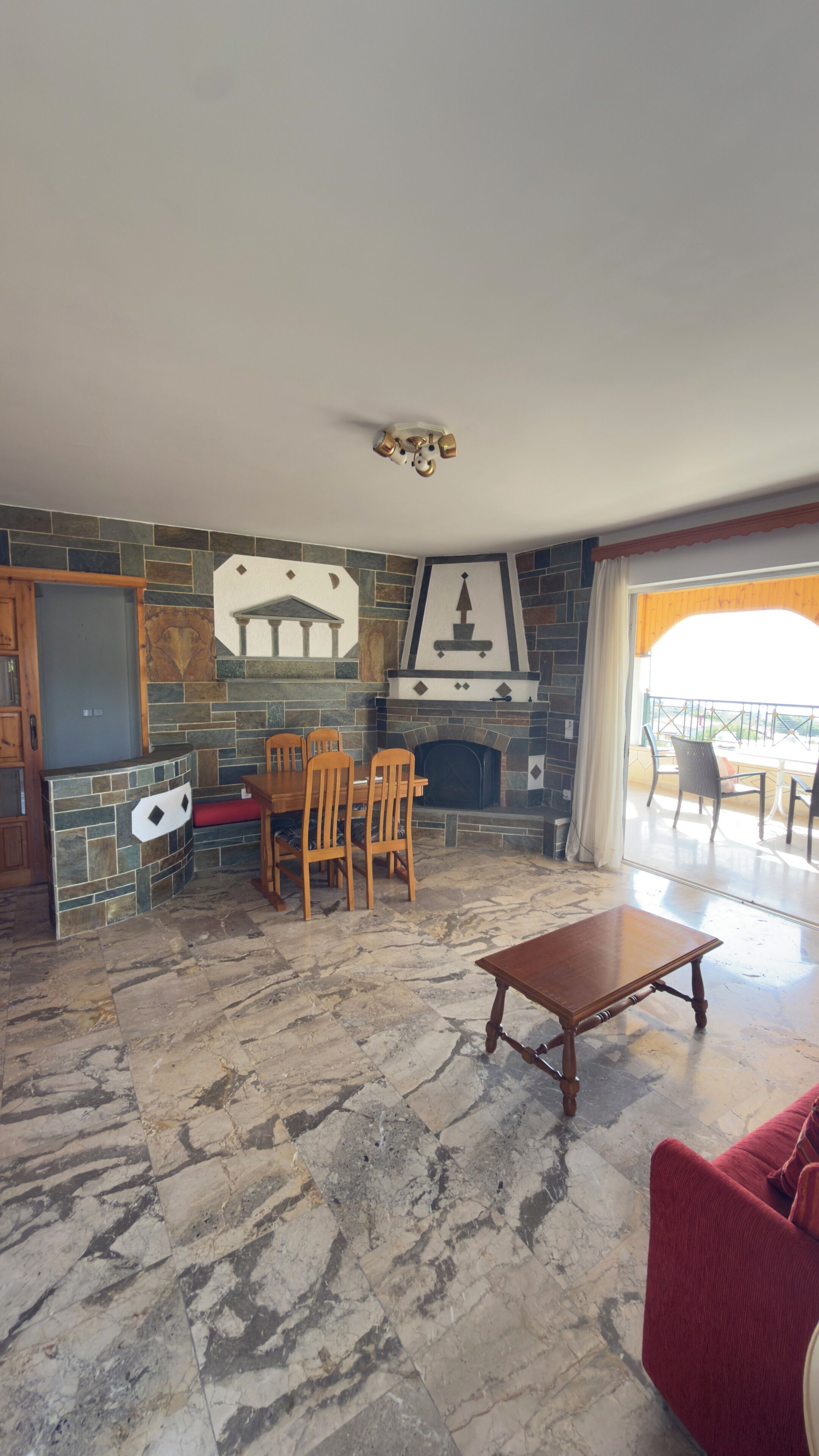 A living room with a stone fireplace, a dining area with chairs, a wooden coffee table, and a view through a glass door.