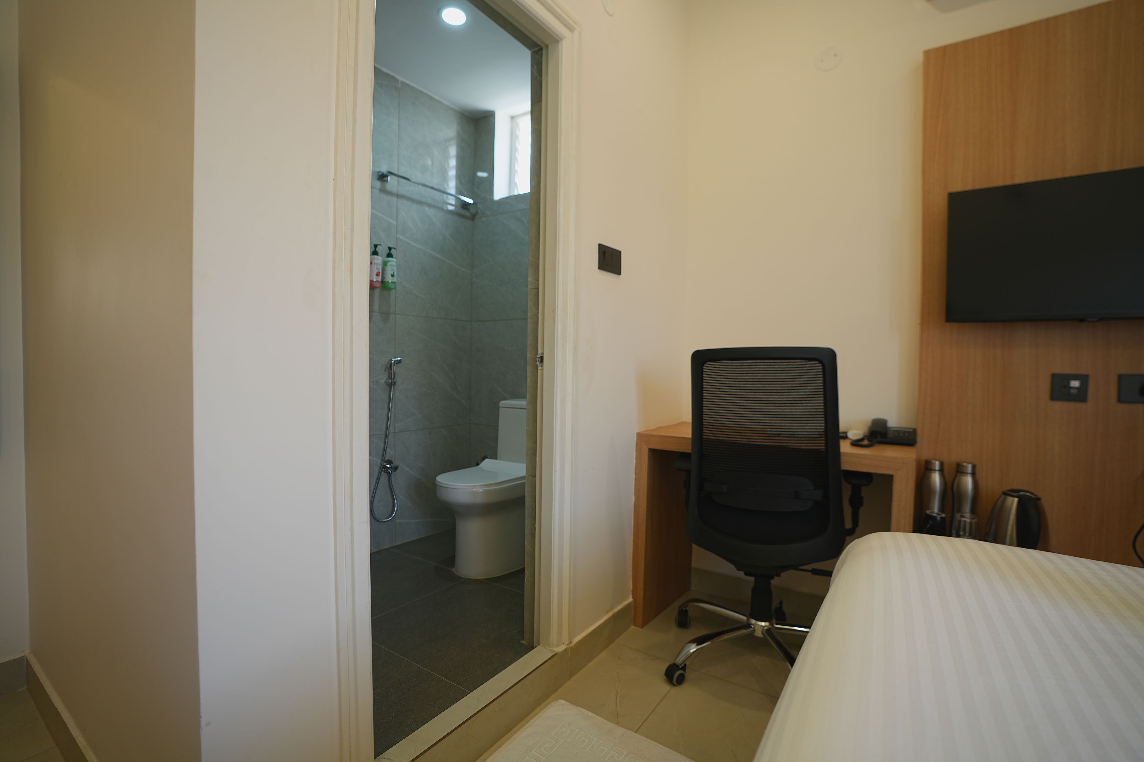 A hotel room with a bed, a desk, a chair, a TV mounted on the wall, and a shower area visible through an open door.