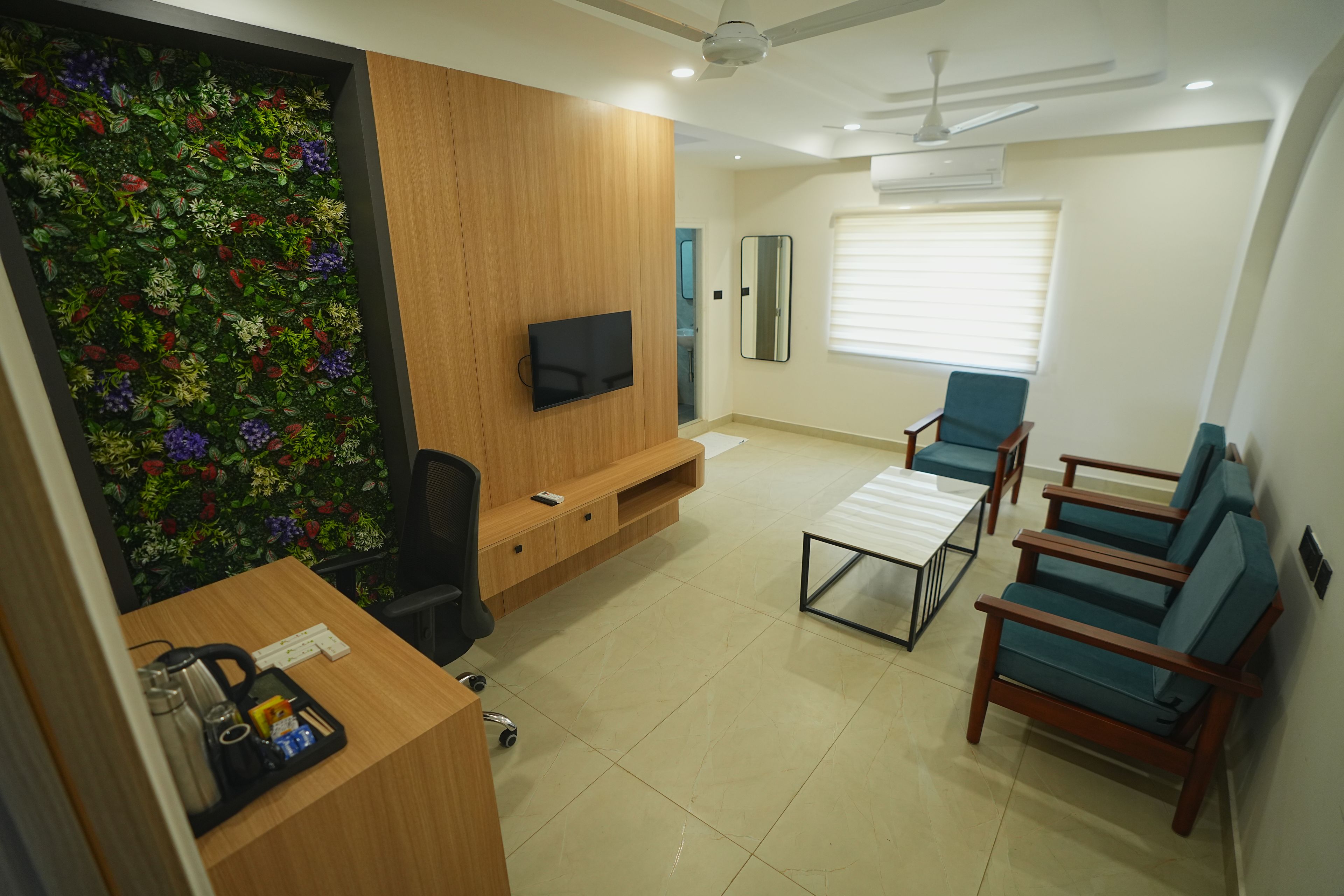 A modern living room with a flat-screen TV, a wooden entertainment unit, a green wall with a floral design, a ceiling fan, and a seating area with blue chairs.