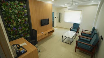 A modern living room with a flat-screen TV, a wooden entertainment unit, a green wall with a floral design, a ceiling fan, and a seating area with blue chairs.