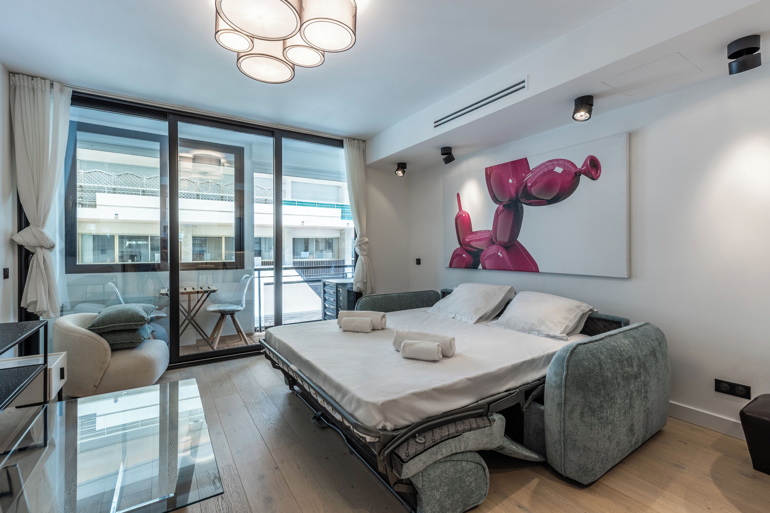 A modern hotel room with a large bed, a seating area with a table and chairs, a large window with a view of buildings, and a unique artwork on the wall.