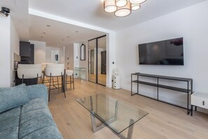 A modern living room with a glass coffee table, a sofa, a TV mounted on the wall, and a kitchen area in the background. - Renovated 1BR | Steps from Croisette & Beach (Cannes)