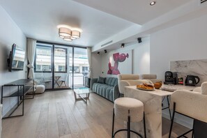 A modern living room with a sofa, coffee table, and bar stools. There is a dining table with breakfast items. The room has large windows and a view of the city. - Renovated 1BR | Steps from Croisette & Beach (Cannes)