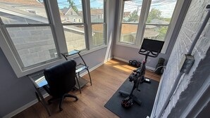 Ein Zimmer mit großen Fenstern, einem Schreibtisch, einem Bürostuhl, einem Heimtrainer und einem Paar Kurzhanteln.