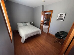 Un dormitorio con cama, mesita de noche, una silla y una puerta que da a una cocina.