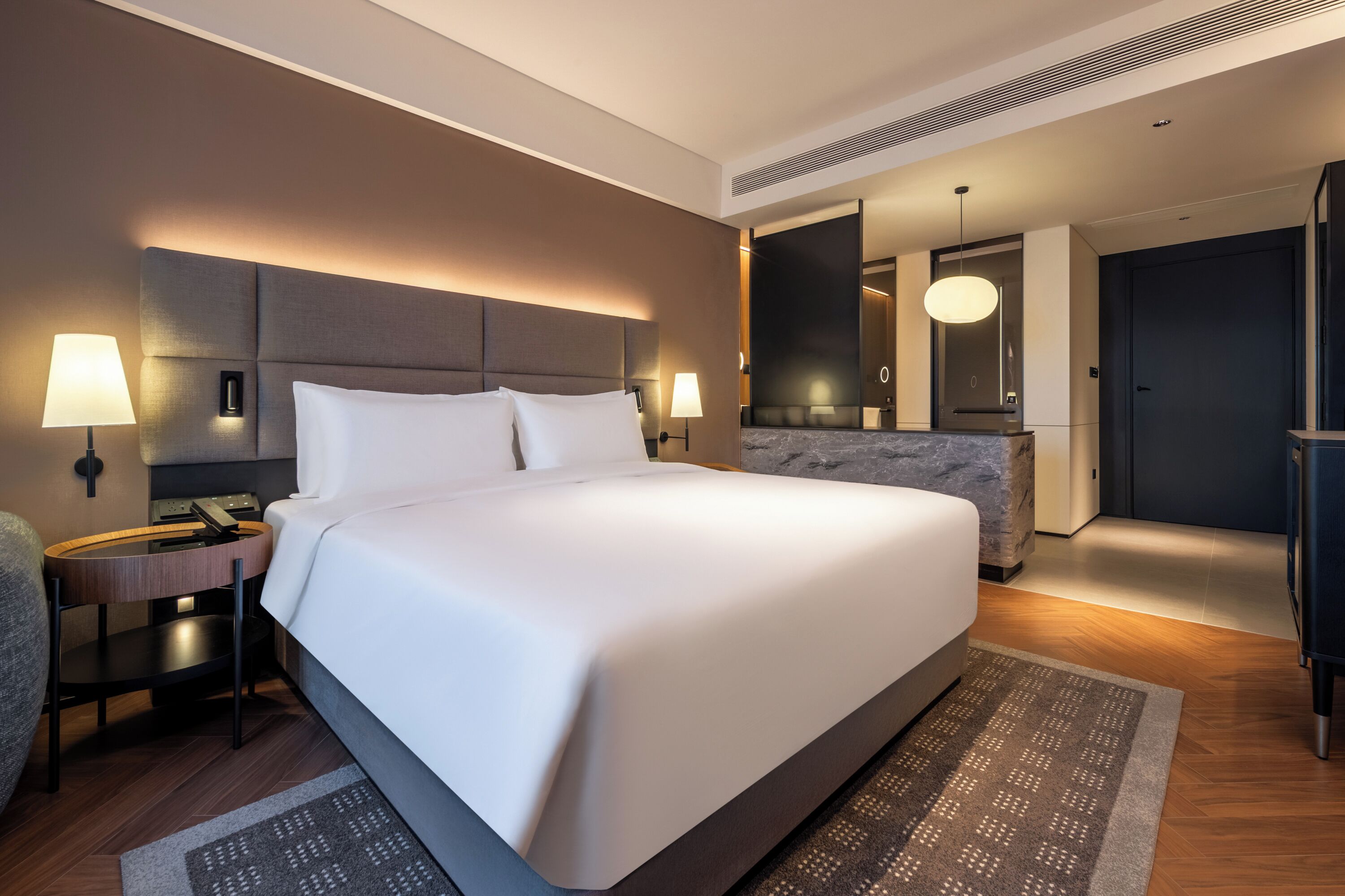 Una habitación de hotel moderna con una cama grande, lámparas de noche, un escritorio y un baño con espejo y toallero.