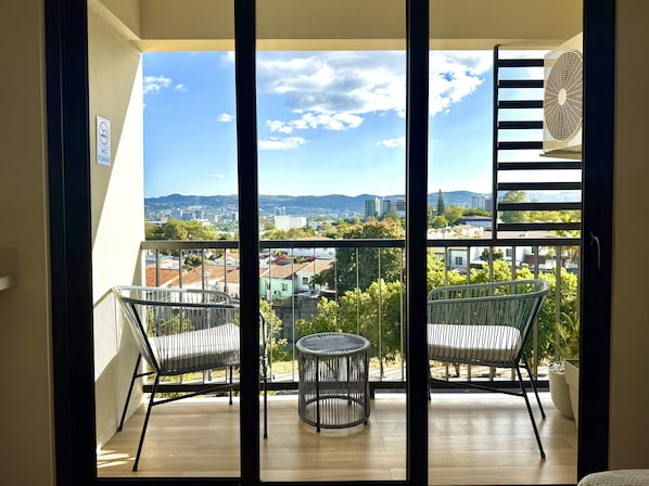 A balcony with two wicker chairs and a small table, overlooking a cityscape with buildings and mountains.