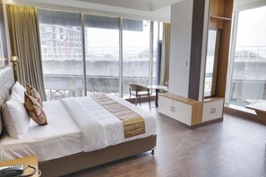 Une chambre d’hôtel avec un grand lit, un bureau, une chaise et une vue sur le paysage urbain grâce à de grandes fenêtres.