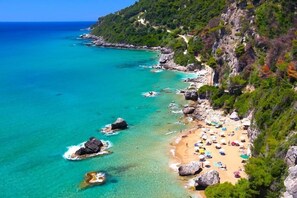 Una platja costanera amb aigües turqueses cristal·lines, roques i una riba sorrenca amb ombrel·les.