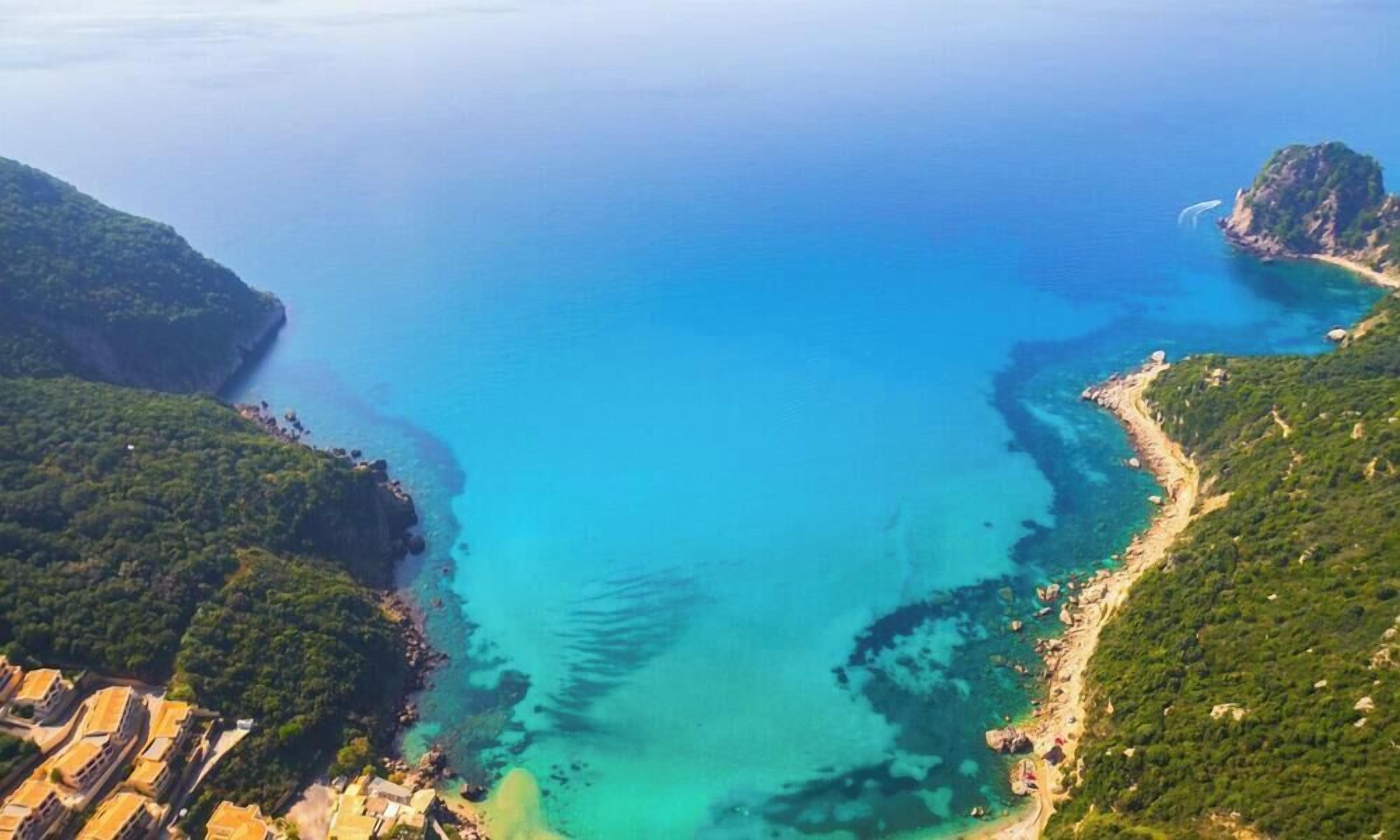 Vista aérea de una zona costera con aguas azules cristalinas, colinas verdes y una pequeña península con edificios.