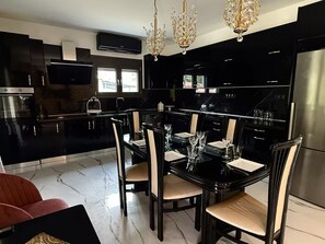 A modern kitchen with a black countertop, stainless steel appliances, and a dining area with a black table and chairs. - Moonlit Olives - Corfu Luxurious & Stylish Escape (Kokkinogia)