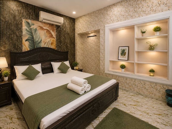 A neatly made bed with a green bedspread and pillows, a large window with shelves displaying plants, and a wall-mounted air conditioner. - Hotel Tejas Inn (Varanasi)