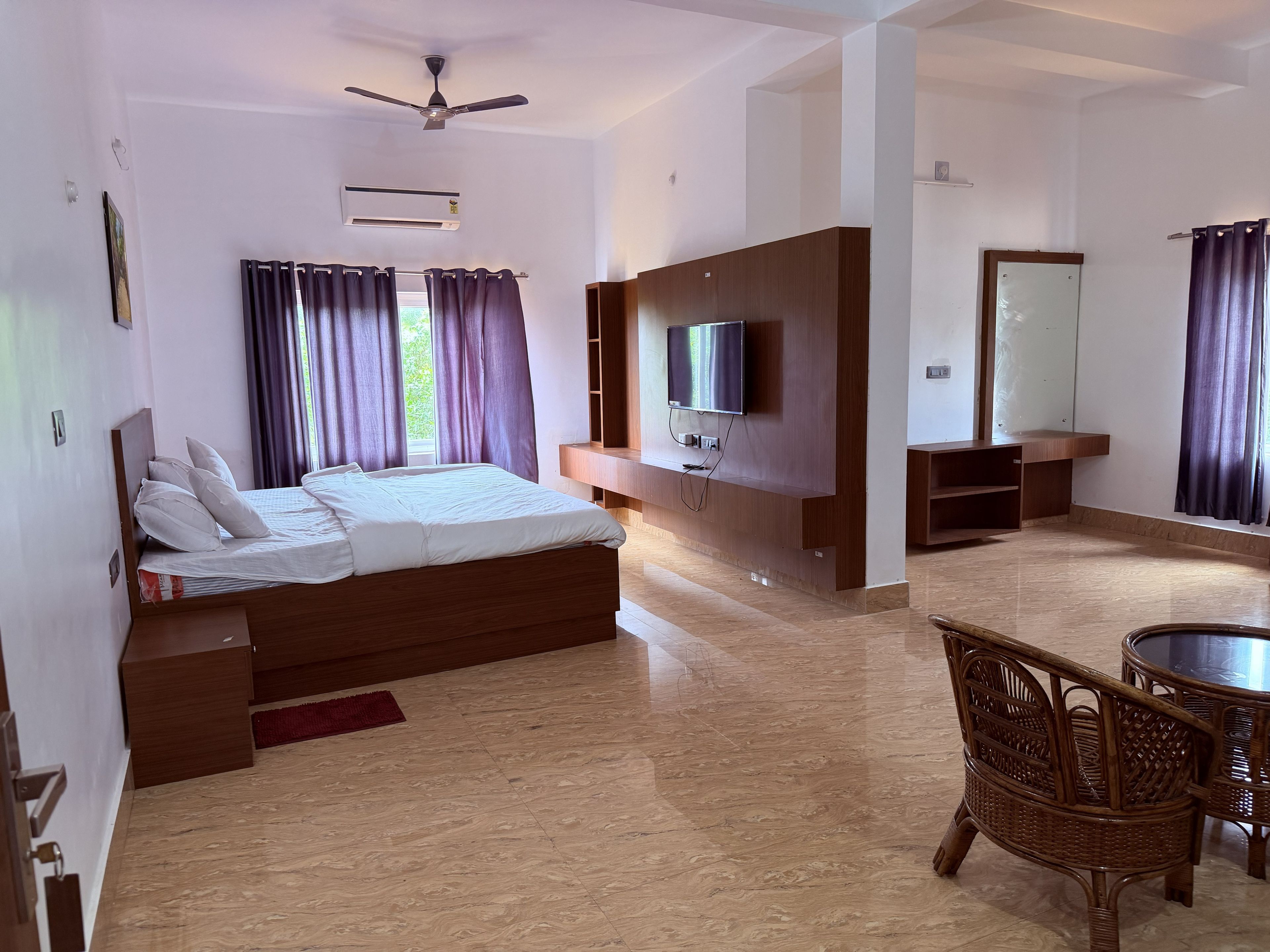 A modern hotel room with a bed, a TV mounted on the wall, a ceiling fan, and a dining area with wicker chairs.