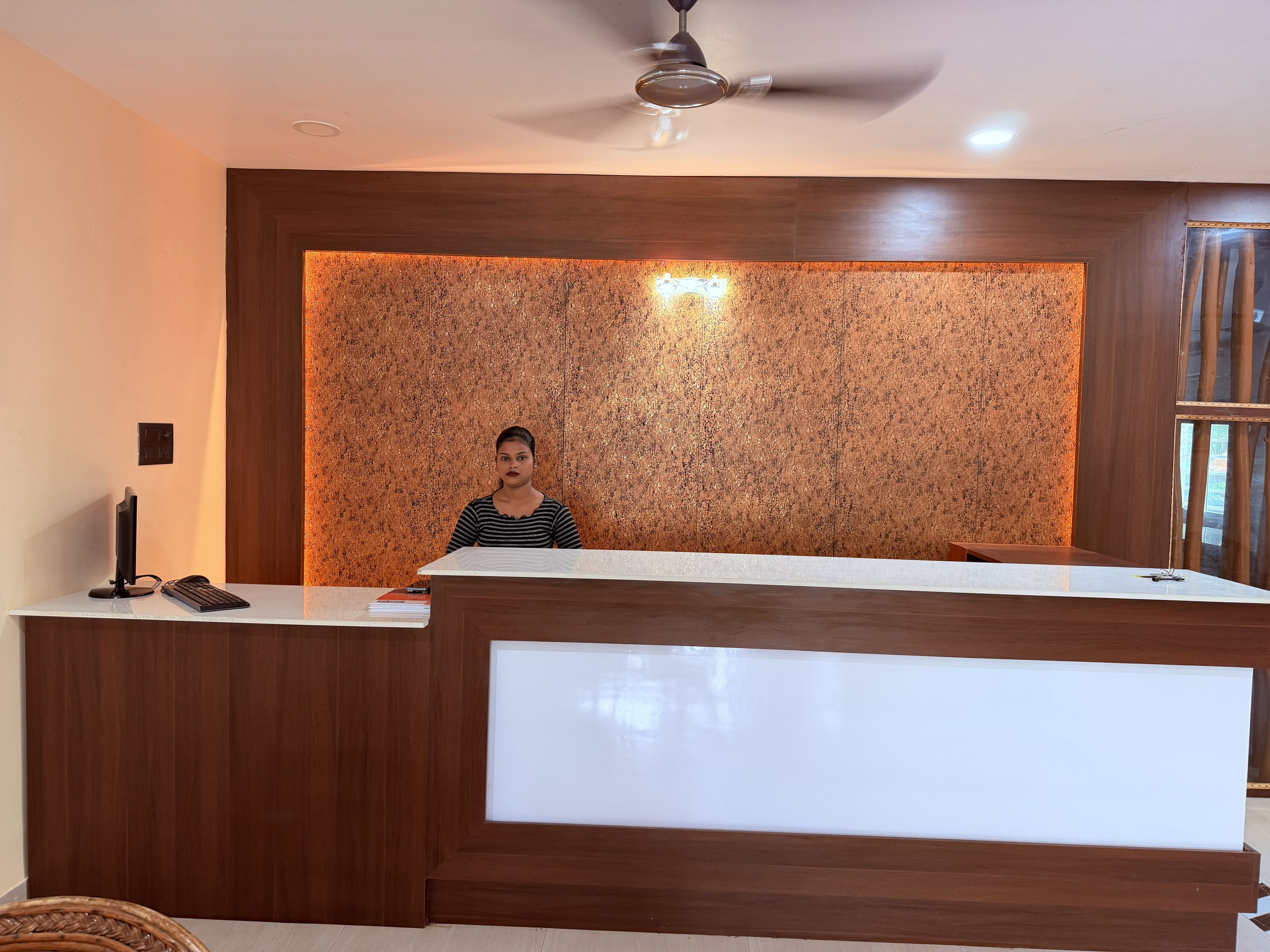 A reception desk with a person standing behind it, a ceiling fan above, and a cork wall behind.