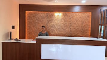 A reception desk with a person standing behind it, a ceiling fan above, and a cork wall behind.