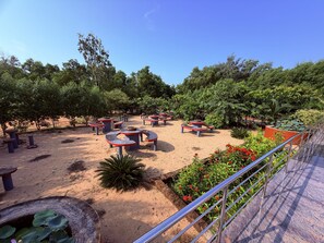 Un espacio al aire libre con mesas circulares y bancos, rodeado de follaje y suelo arenoso.
