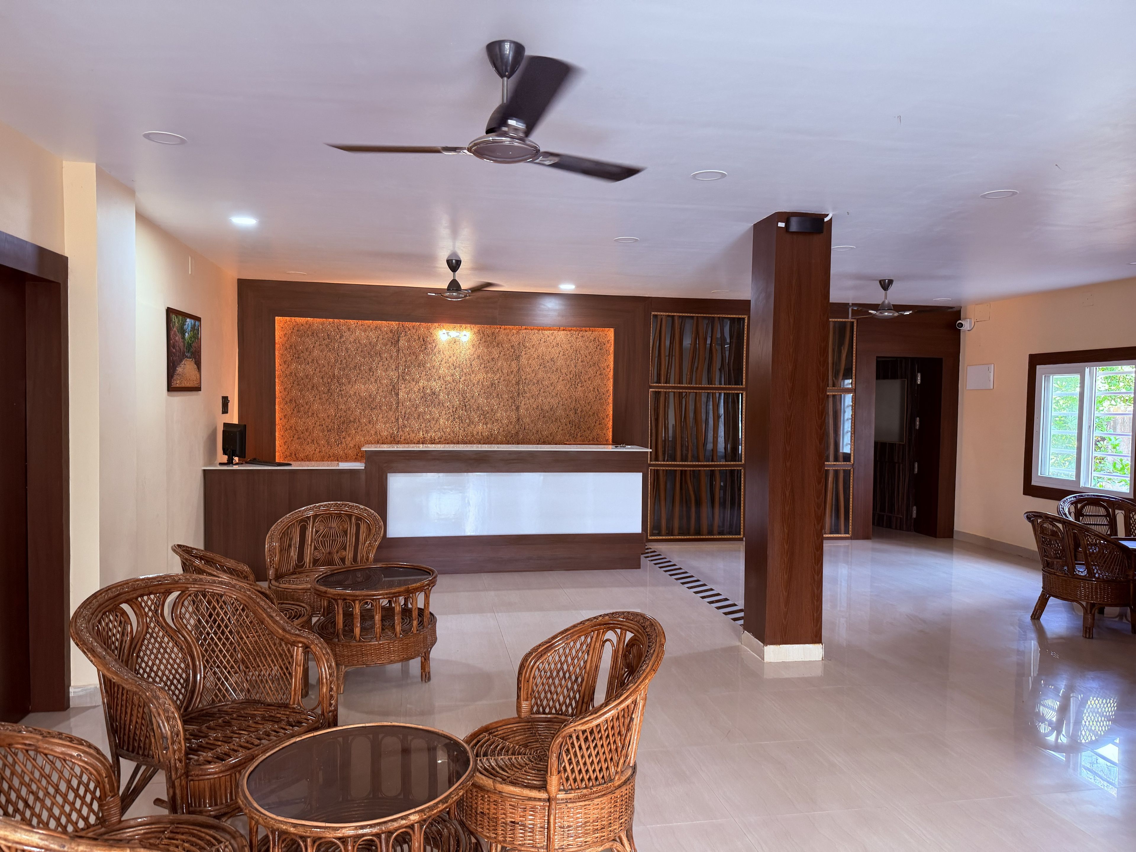 A hotel lobby with a reception desk, wicker furniture, a ceiling fan, and a large window.