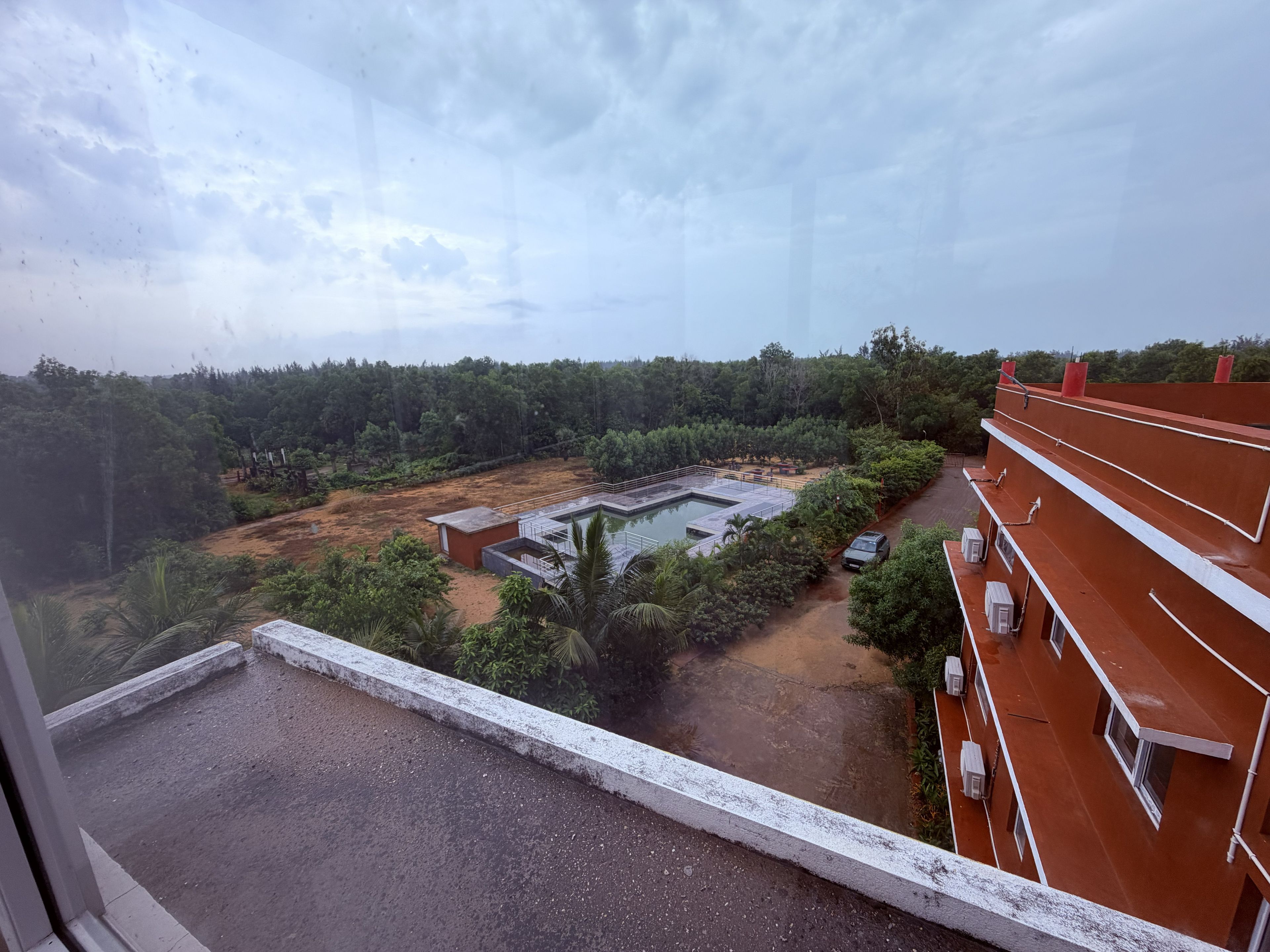 Vista da janela sobre um pátio com piscina, rodeado por árvores e um edifício com fachada vermelha.
