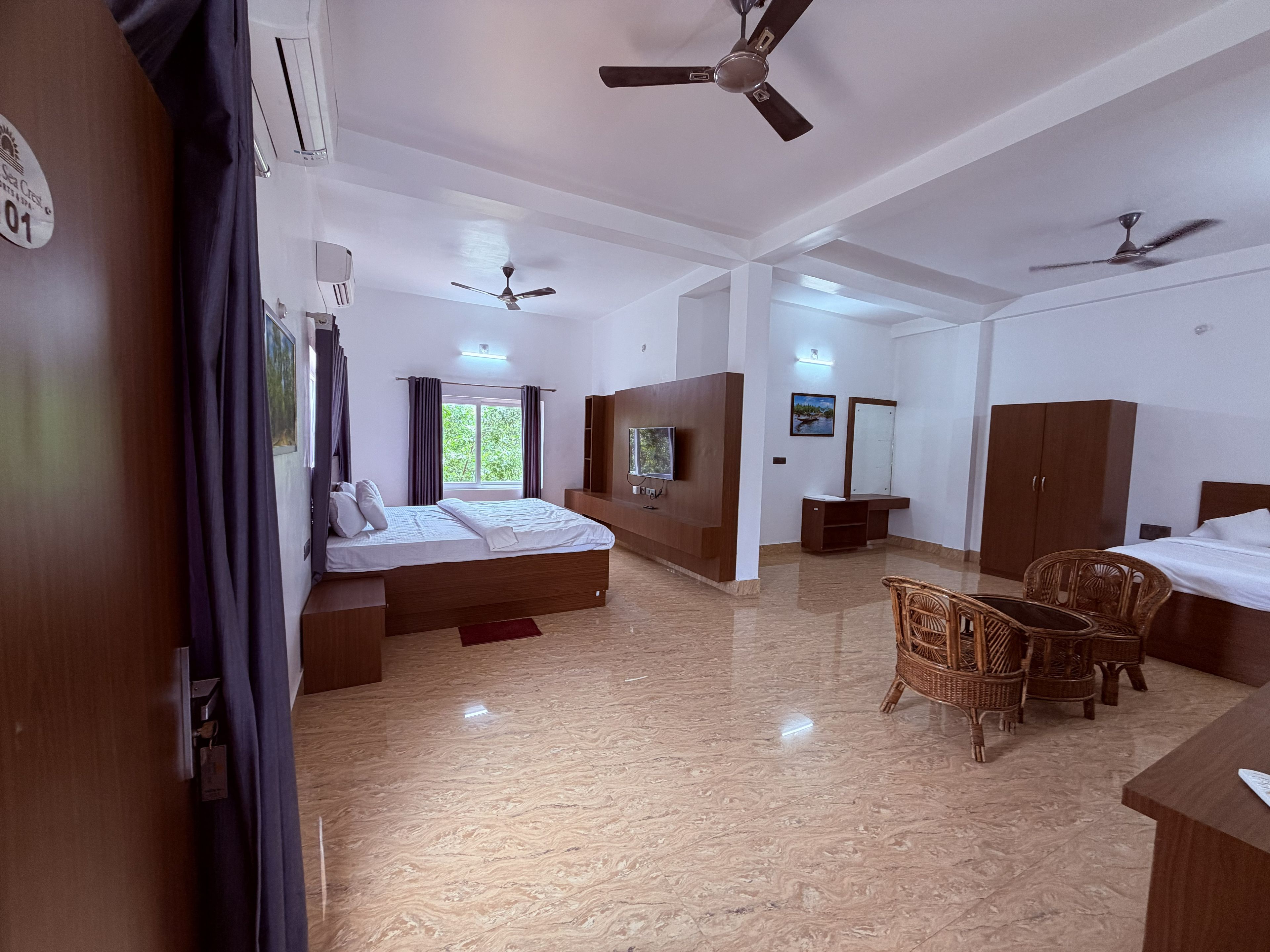 A hotel room with a bed, ceiling fans, a TV, a wardrobe, and a seating area with chairs.