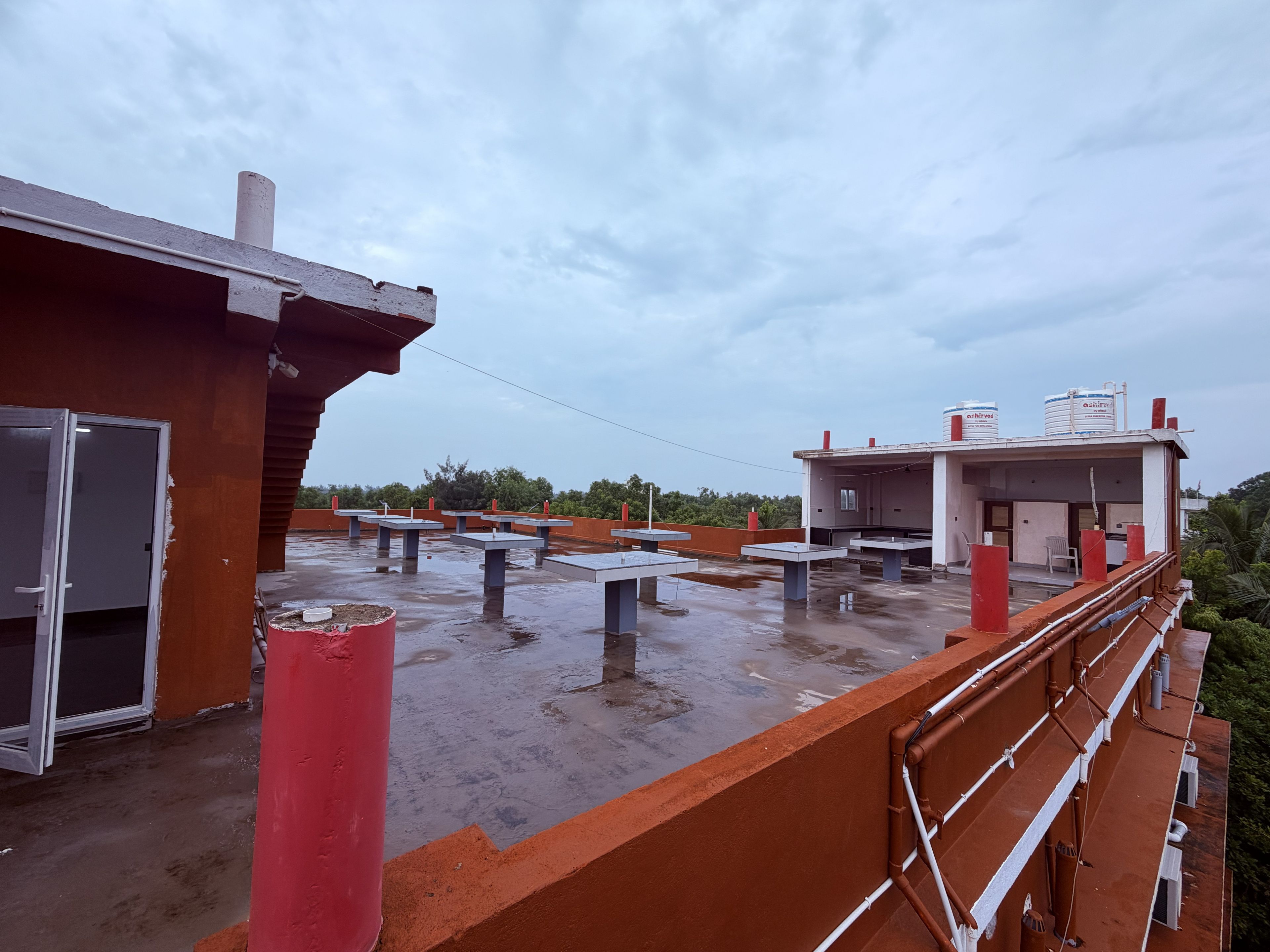 Una terraza en la azotea con varias mesas y sillas, un tanque de agua y un edificio con decoración roja y blanca.