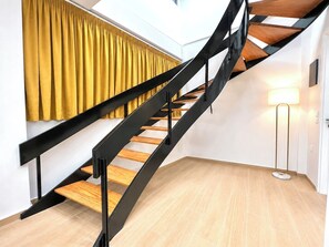 A modern staircase with black metal railings and wooden steps, leading to a room with a yellow curtain and a floor lamp. - Galleria The House by ΜΡS (Athina)