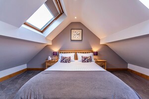 Un dormitorio con techo inclinado, tragaluz, cama con almohadas, mesitas de noche y un cuadro enmarcado en la pared.