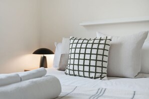 Ein ordentlich bezogenes Bett mit weißen Leinen und einem karierten Kopfkissen, eine Nachttischlampe und eine weiße Wand.