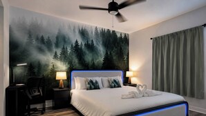 Ein Schlafzimmer mit einem Bett, einem Schreibtisch, einem Sessel, einem Deckenventilator und einem Wandbild von einem Wald.