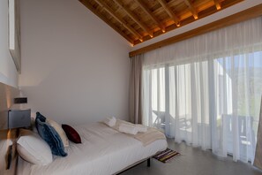 Ein Schlafzimmer mit einem großen Bett, einer Holzbalkendecke und Blick ins Freie durch transparente Vorhänge.