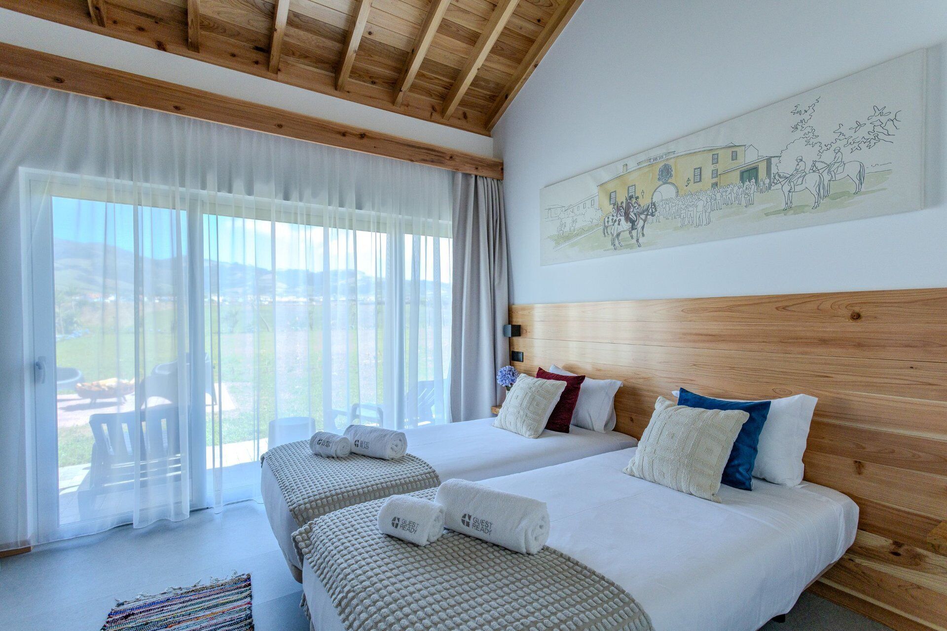 A hotel room with two beds, a wooden ceiling, a large window with curtains, and a wall painting.