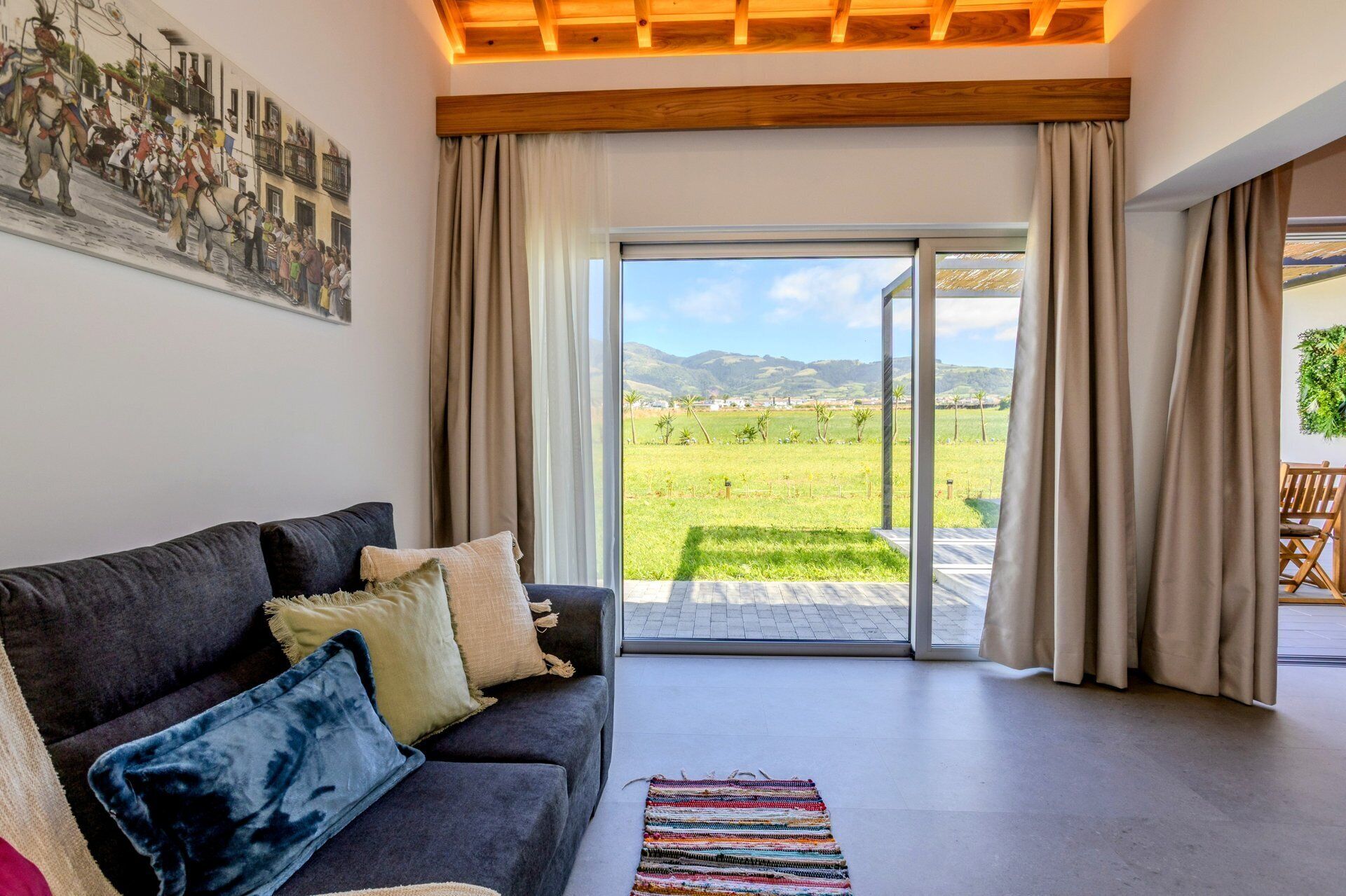 Een woonkamer met een bank, een vloerkleed en uitzicht op een groen veld door een glazen deur.