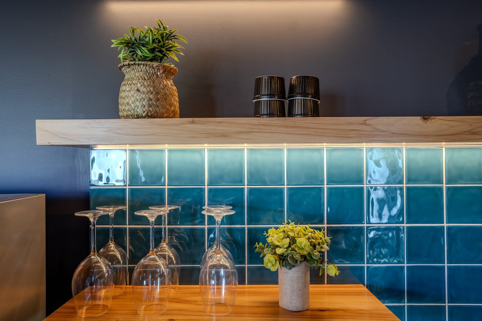 A kitchen area with a wooden shelf, a blue tiled backsplash, and a wooden table with wine glasses and a vase with green plants.