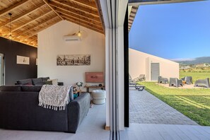 Un intérieur moderne avec un canapé foncé, un tableau et un panneau indiquant « PRÊT POUR LES INVITÉS ». À l’extérieur, on trouve une terrasse avec des chaises et une vue sur la campagne.
