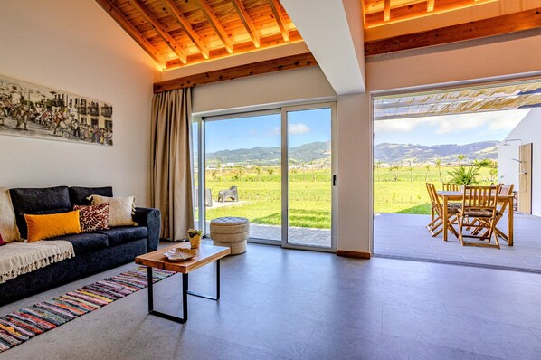 A modern living room with a sofa, a coffee table, and a view of a green landscape through large glass doors.