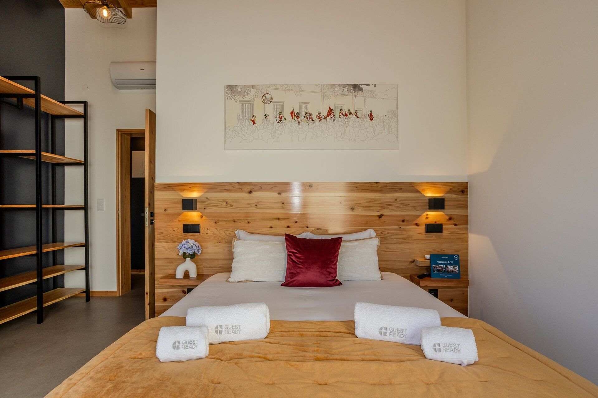 A hotel room with a wooden headboard, a bed with white and red pillows, and a wall-mounted artwork depicting a historical scene.
