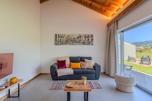A modern living room with a dark sofa, a flat-screen TV, a coffee table, and a large window offering a view of the outdoors. - GuestReady - Casa do Cavaleiro by Cavalhadas House (Ribeira Grande)