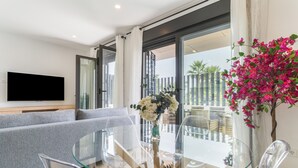 Een moderne woonkamer met een glazen eettafel, een grijze bank en een flatscreen televisie aan de muur.